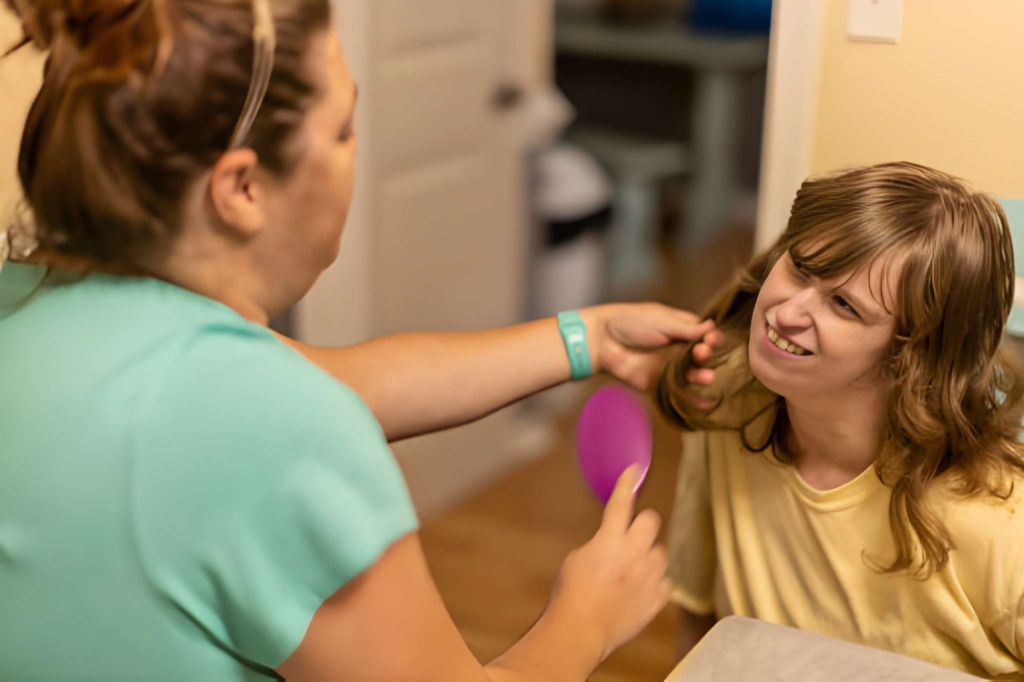 Caregiver in green scrubs gently brushes a young woman's hair with a pink brush, both smiling in a clinic room.