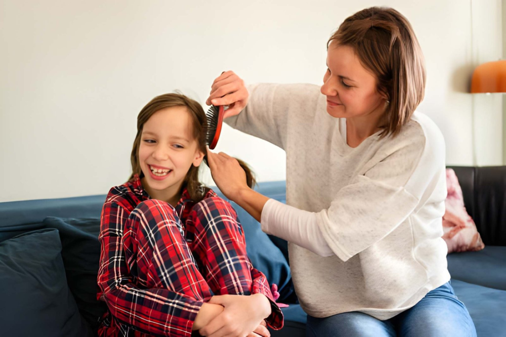 Woman combing a young girl's hair as they sit on a blue couch; girl in red plaid pajamas smiles.