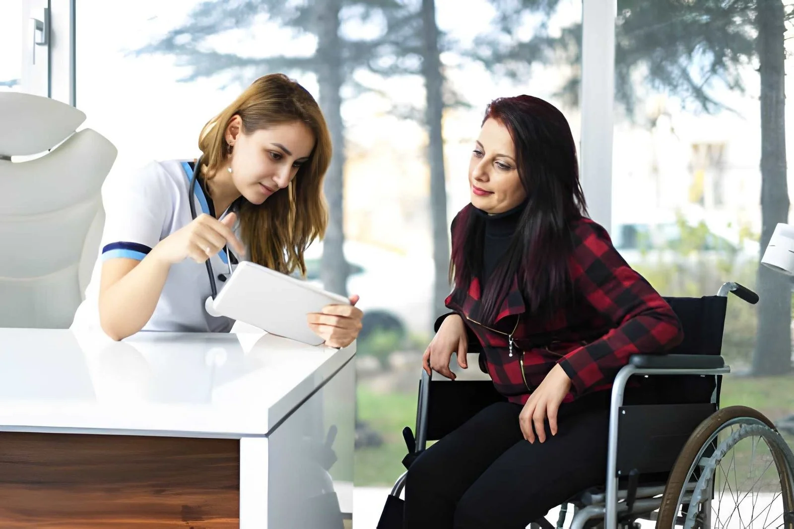 Nurse or healthcare worker shows a tablet to a woman seated in a wheelchair at a clinic reception desk, discussing something.